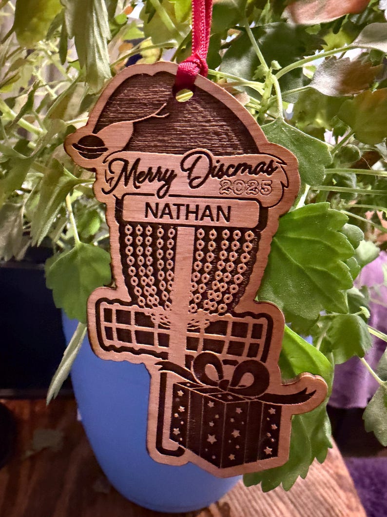 K&ouml;nnte beinhalten: Weihnachtsbaumschmuck aus Holz mit einem Disc-Golf-Korb-Design. Der Schmuck tr&auml;gt den Text "Merry Discmas 2025" und den Namen "Nathan". Ein rotes Band ist zum Aufh&auml;ngen befestigt. Der Schmuck ist braun mit einem Geschenkbox-Design.