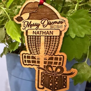 K&ouml;nnte beinhalten: Weihnachtsbaumschmuck aus Holz mit einem Disc-Golf-Korb-Design, einem Weihnachtsmannhut und einer Geschenkbox. Der Schmuck ist mit "Merry Discmas 2025" und dem Namen "Nathan" graviert. Ein rotes Band ist zum Aufh&auml;ngen befestigt.