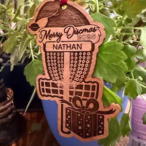 K&ouml;nnte beinhalten: Weihnachtsschmuck aus Holz mit Disc-Golf-Motiv. Der Schmuck ist wie ein Disc-Golf-Korb mit Weihnachtsm&uuml;tze und Geschenkbox geformt. Der Text "Merry Discmas 2025" und "Nathan" sind eingraviert. Ein rotes Band ist zum Aufh&auml;ngen befestigt.