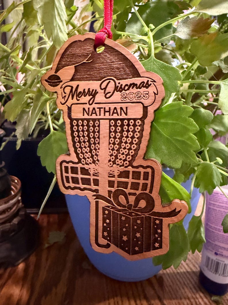 K&ouml;nnte beinhalten: Weihnachtsschmuck aus Holz mit Disc-Golf-Motiv. Der Schmuck ist wie ein Disc-Golf-Korb mit Weihnachtsm&uuml;tze und Geschenkbox geformt. Der Text "Merry Discmas 2025" und "Nathan" sind eingraviert. Ein rotes Band ist zum Aufh&auml;ngen befestigt.
