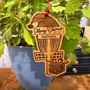 K&ouml;nnte beinhalten: Weihnachtsbaumschmuck aus Holz f&uuml;r Disc Golf mit rotem Band. Der Schmuck zeigt ein Disc Golfkorb-Design mit dem Text "Merry Discmas 2025" und dem Namen "Nathan". Eine Geschenkbox ist ebenfalls enthalten.