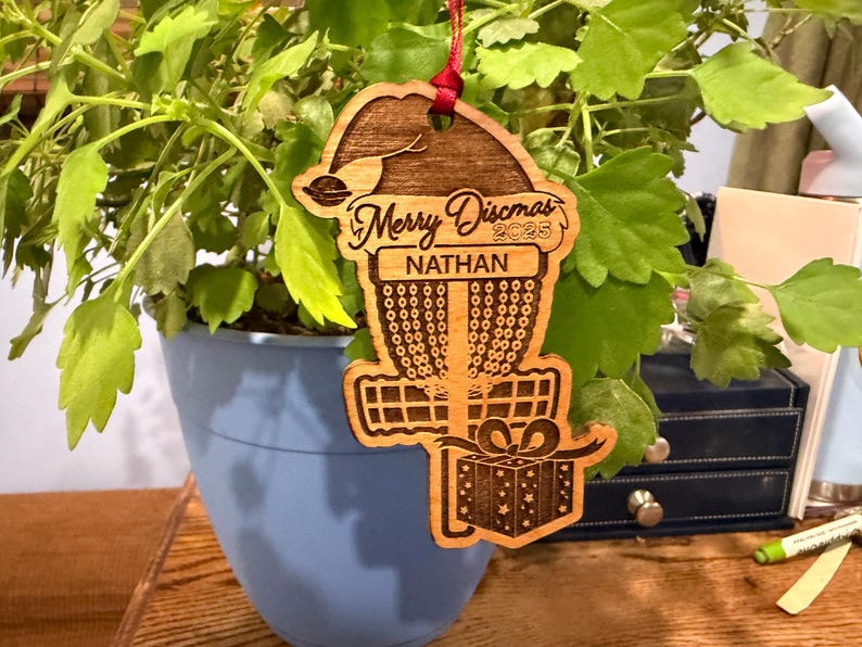 K&ouml;nnte beinhalten: Weihnachtsbaumschmuck aus Holz f&uuml;r Disc Golf mit rotem Band. Der Schmuck zeigt ein Disc Golfkorb-Design mit dem Text "Merry Discmas 2025" und dem Namen "Nathan". Eine Geschenkbox ist ebenfalls enthalten.