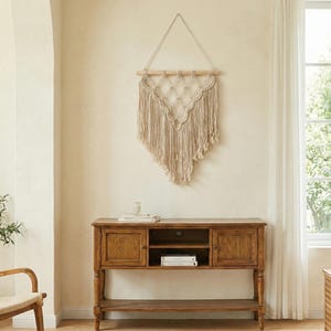 Könnte beinhalten: Ein beigefarbener Makramee-Wandbehang mit geometrischem Design und Fransen hängt über einer hölzernen Konsolentisch. Der Tisch hat zwei Türen, ein offenes Regal und ein unteres Regal. Der Raum hat eine neutrale Farbpalette mit natürlichem Licht.