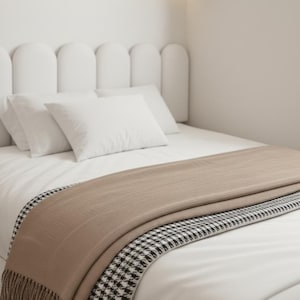 May include: A bed with a white headboard, white pillows, and a white comforter. A tan throw blanket with a black and white houndstooth border is draped across the bed. The throw has a fringed edge.