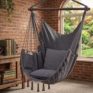 May include: A dark grey hanging chair with a matching cushion and tassels. The chair is suspended from a wooden bar and rope, with pockets on the side. The background features a brick wall and a window.
