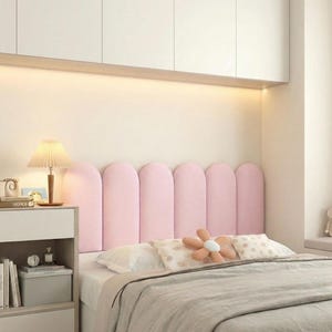 May include: A bedroom with a pink, arched headboard. The bed is made with a grey duvet and white pillows. A flower-shaped cushion and a bedside table with a lamp complete the scene. The room has a neutral colour palette.
