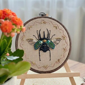 Può includere: Un'opera d'arte ricamata a mano su telaio, raffigurante un dettagliato scarabeo. Lo scarabeo è impreziosito da perline verdi, nere e dorate. L'opera è esposta su un piccolo cavalletto di legno.