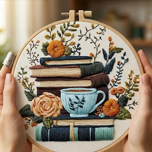 May include: Embroidered hoop art with a stack of books, a teacup, and floral details in brown, blue, orange, and green. The design is set against a cream background, creating a cosy, handcrafted aesthetic. The hoop is approximately 20 cm in diameter.