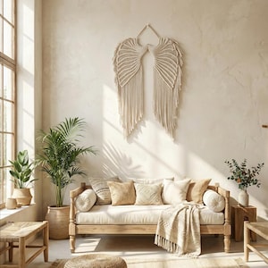 Op de afbeelding: Een neutrale woonkamer met een macramé engelenvleugel wanddecoratie. Een licht houten daybed is versierd met kussens en een franjedeken. Potplanten voegen een natuurlijk tintje toe. Een geweven poef en vloerkleed maken de gezellige sfeer compleet.
