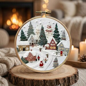 May include: An embroidered winter village scene in a wooden hoop. The artwork shows snow-covered houses, fir trees, and figures. The hoop is displayed on a wooden slice, creating a festive atmosphere. The scene is reminiscent of a Christmas card.