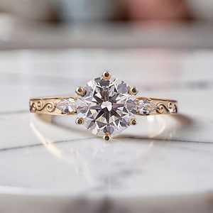 Könnte beinhalten: Ein goldener Verlobungsring mit einem großen, runden, klaren Edelstein als Mittelpunkt. Zwei kleinere tropfenförmige Steine flankieren den Hauptstein. Das Band weist ein verziertes, wirbelndes Design auf.
