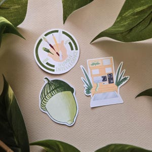 May include: Three stickers on a beige surface. One sticker is a circle with the text "MADE WITH HUMAN INTELLIGENCE" and a hand holding art supplies. Another is an acorn. The third is a yellow and white vending machine.