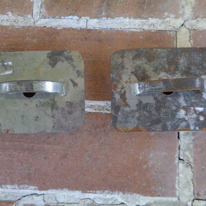 May include: Two weathered, rectangular metal objects with a handle and a small hole. The objects are silver and have a worn, aged appearance. They are set against a red brick wall.