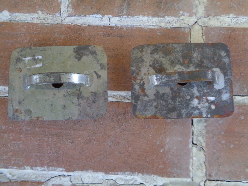 May include: Two weathered, rectangular metal objects with a handle and a small hole. The objects are silver and have a worn, aged appearance. They are set against a red brick wall.