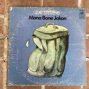 May include: A vintage vinyl record album cover featuring a blue background with a green and white illustration of a trash can. The album title "Mona Bone Jakon" is printed in white text above the illustration. The artist's name "Cat Stevens" is printed in white text above the album title.