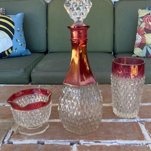 Può includere: Una caraffa di vetro trasparente con collo e tappo colorati di rosso, insieme a un bicchiere e una crema abbinati. La caraffa presenta un motivo a taglio diamante e il tappo è di cristallo sfaccettato. Il bicchiere e la crema hanno un bordo rosso.