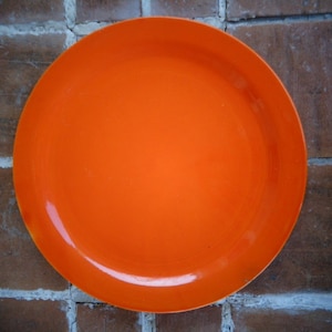 Könnte beinhalten: Ein leuchtend orangefarbener Teller mit leicht erhöhtem Rand. Der Teller ist rund und scheint aus glänzendem Material zu bestehen. Der Teller liegt auf einer Ziegeloberfläche.