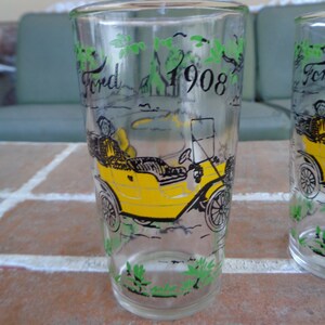 May include: Two clear glass drinking glasses with a yellow car and the text "Ford 1908" printed on them. The glasses have green leaves around the top and bottom.