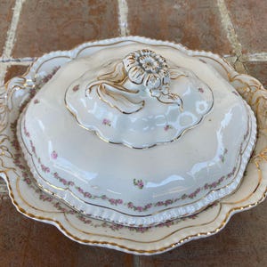 May include: A white porcelain butter dish with a lid. The dish is decorated with a delicate floral pattern in pink and gold. The lid has a raised floral design in the center.
