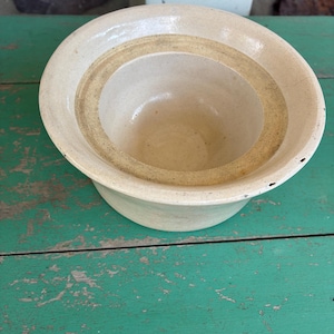 May include: A round, off-white ceramic bowl with a wide, flat rim and a deep, circular basin. The interior features a distinct, slightly darker beige ring around the center, and the exterior surface shows a smooth, glazed finish with minor imperfections.