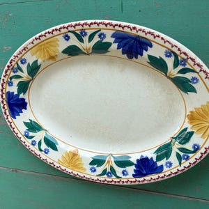 May include: An oval ceramic serving platter featuring a cream-colored center and a decorative border. The rim is adorned with a repeating pattern of yellow and blue stylized flowers, green leaves, and small blue blossoms, finished with a thin red edge.