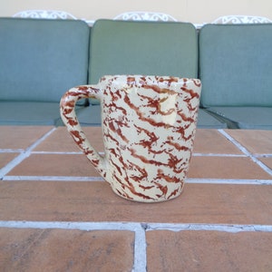 Può includere: Una tazza in ceramica con base color crema e un motivo astratto marrone. La tazza ha un manico grande ed è appoggiata su una superficie di mattoni. Lo sfondo comprende cuscini verdi e un telaio in metallo bianco.