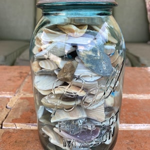 May include: A clear glass jar filled with various seashells. The jar has a blue-green tint and a silver lid. The words "Perfect Mason" are embossed on the jar. The seashells are beige, white, and gray.