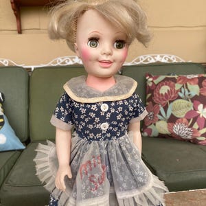 May include: A doll with blonde hair styled with a white ribbon, green eyes, and rosy cheeks. The doll wears a navy blue dress with a small white floral pattern, a rounded collar, two white buttons, and a sheer white ruffled apron with the text Miss Sue.
