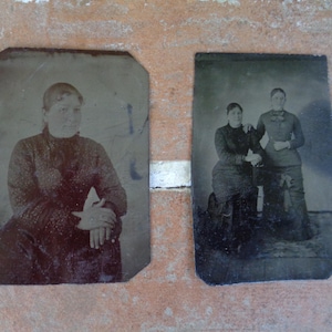 May include: Two vintage black and white tintype photographs of women. The first photo shows a woman wearing a dark patterned dress and sitting with her hands folded in her lap. The second photo shows two women standing side-by-side. The woman on the left is wearing a dark dress and the woman on the right is wearing a dark jacket and skirt.