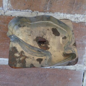May include: A vintage, rectangular cookie cutter with a bird-like design. The metal cutter has a tarnished, aged appearance with rust spots. The cutter is set against a background of red bricks.