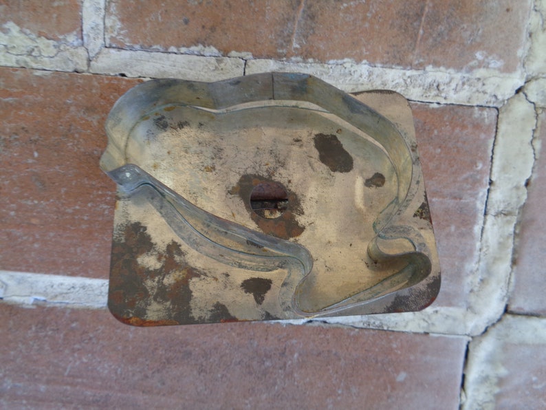 May include: A vintage, rectangular cookie cutter with a bird-like design. The metal cutter has a tarnished, aged appearance with rust spots. The cutter is set against a background of red bricks.