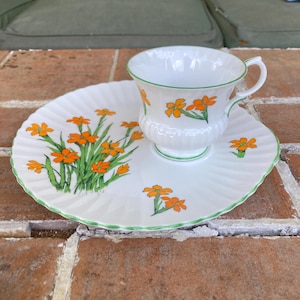 Puede incluir: Juego de taza y platillo blancos con diseño floral naranja y ribete verde. El platillo tiene un borde festoneado y un patrón floral a juego. La taza tiene un asa y un diseño floral similar. El juego está sobre una superficie de ladrillo.