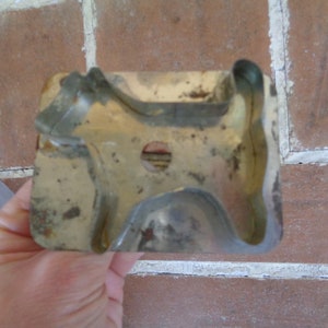 May include: Vintage metal dog-shaped cookie cutter. The cutter has a rectangular base with a dog silhouette cut out. The metal has a worn, aged appearance with some rust spots, suggesting it's an antique kitchen tool.