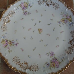 Puede incluir: Un plato de servicio de porcelana blanca con un borde dorado y un diseño floral. El plato presenta un delicado patrón de pequeñas flores azules y rosas, así como un motivo floral más grande en el centro.