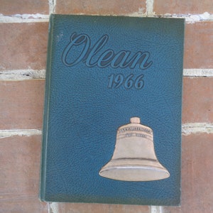 May include: A dark blue hardcover book with the title "Olean 1966" embossed on the front cover. A silver bell with the inscription "Olean Valley School" is also on the cover.