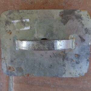 May include: A rectangular, silver-colored metal object with a curved handle. The metal has a worn, aged appearance with some rust spots. The handle is attached to the top of the rectangle. The object appears to be a tool or part of a larger mechanism.