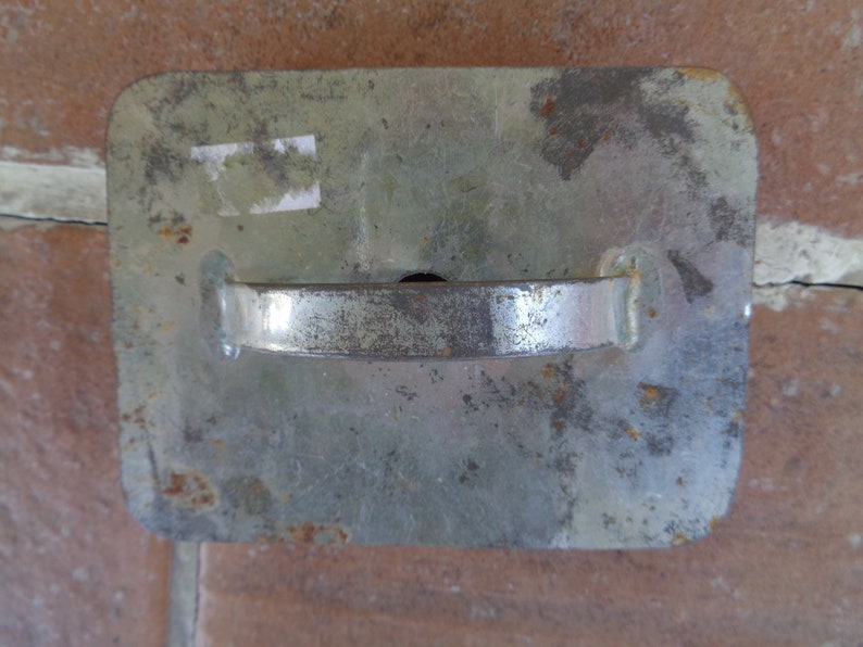 May include: A rectangular, silver-colored metal object with a curved handle. The metal has a worn, aged appearance with some rust spots. The handle is attached to the top of the rectangle. The object appears to be a tool or part of a larger mechanism.