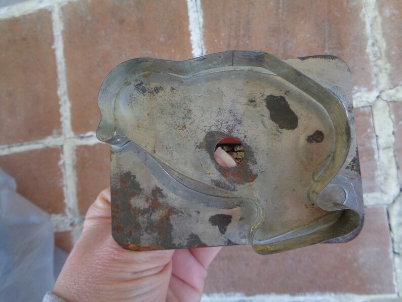 May include: A vintage, rectangular cookie cutter with a heart-shaped design. The metal cutter has a worn, aged appearance with areas of rust and discoloration. The cutter has a small hole in the center.