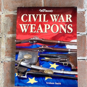 May include: A book titled "Warman's Civil War Weapons" with a red cover and white text. The cover features images of rifles, pistols, and other weaponry. A blue flag with yellow stars is visible.