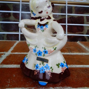 May include: A white ceramic figurine of a girl wearing a blue and brown dress with a blue flower pattern. The figurine is holding a bouquet of flowers and has the word "NINA" written on a blue tag.