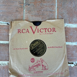 May include: A vintage vinyl record inside a tan paper sleeve featuring the RCA Victor logo in red text. The center label is black with gold text reading Decca, The Anniversary Waltz, and Guy Lombardo. A small dog and gramophone icon is printed below.