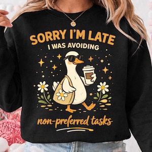 May include: Black sweatshirt with a graphic of a goose wearing sunglasses and holding a coffee cup and tote bag. Text reads "SORRY I'M LATE I WAS AVOIDING non-preferred tasks". The design includes flowers and stars.