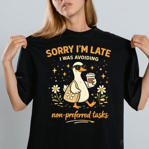 May include: Black t-shirt with a graphic of a duck wearing sunglasses, carrying a tote bag and coffee cup. Text reads "SORRY I'M LATE I WAS AVOIDING non-preferred tasks". The duck is surrounded by stars and flowers.