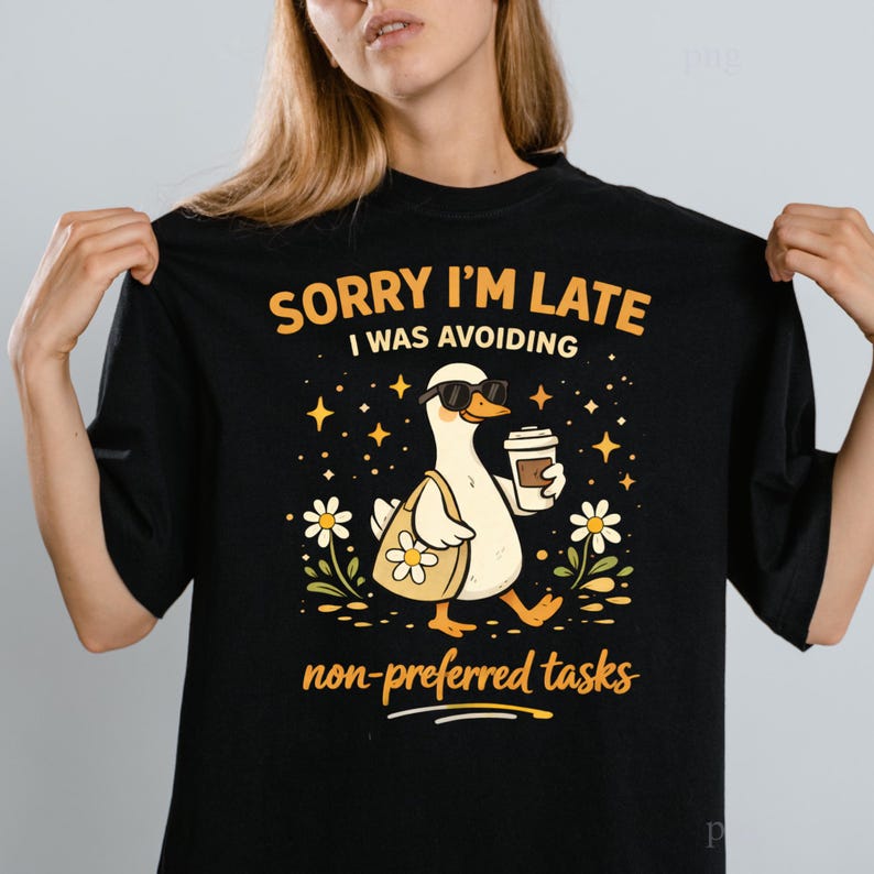 May include: Black t-shirt with a graphic of a duck wearing sunglasses, carrying a tote bag and coffee cup. Text reads "SORRY I'M LATE I WAS AVOIDING non-preferred tasks". The duck is surrounded by stars and flowers.