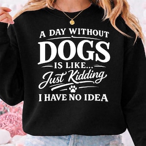 Puede incluir: Sudadera negra con texto blanco: "A DAY WITHOUT DOGS IS LIKE... Just Kidding I HAVE NO IDEA" y una huella de pata. La sudadera es de cuello redondo.