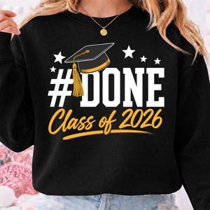 Può includere: Felpa nera con la scritta "#DONE Class of 2026" in bianco e oro. Una grafica del cappello di laurea è sopra la parola "DONE", con stelle. La felpa è a girocollo.