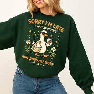 May include: Dark green sweatshirt with the text "SORRY I'M LATE I WAS AVOIDING non-preferred tasks". The graphic features a cartoon duck wearing sunglasses, carrying a tote bag and a coffee cup, surrounded by flowers and stars.
