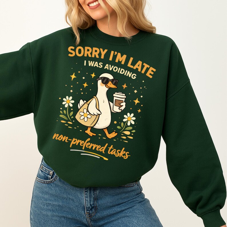 May include: Dark green sweatshirt with the text "SORRY I'M LATE I WAS AVOIDING non-preferred tasks". The graphic features a cartoon duck wearing sunglasses, carrying a tote bag and a coffee cup, surrounded by flowers and stars.