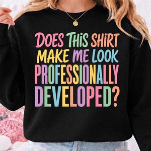 Pode incluir: Uma camisola preta de manga comprida com o texto colorido "DOES THIS SHIRT MAKE ME LOOK PROFESSIONALLY DEVELOPED?" impresso em tons de rosa, verde, laranja, amarelo e azul na zona do peito da peça de vestuário.