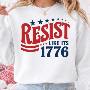 Op de afbeelding: Een wit sweatshirt met ronde hals en een grafische print in rood en marineblauw. De tekst luidt RESIST LIKE ITS 1776, vergezeld door een rij marineblauwe sterren boven het woord RESIST en drie kleine rode sterren bij de tekst.
